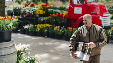 Male UPS driver carrying two packages down a flower lined sidewalk