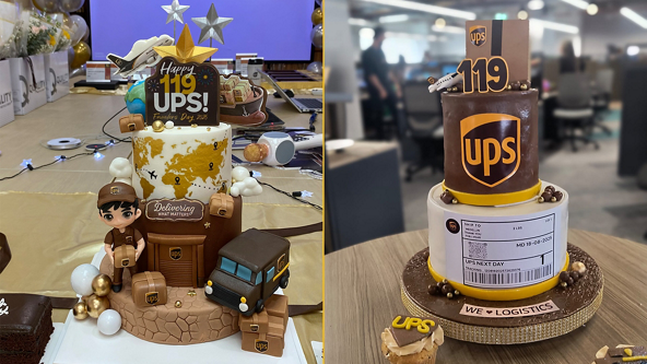 Two elaborately decorated cakes celebrate the 119th anniversary of UPS in an office setting. The left cake features a world map design, a small UPS delivery truck, parcels, and a cartoon delivery worker figurine surrounded by gold and white balloons. The right cake is topped with a large UPS shield, the number 119, and a tier styled like a shipping label, sitting on a base with the phrase "WE ♥ LOGISTICS". Both cakes prominently display the UPS name and colors, emphasizing global delivery and logistics.
