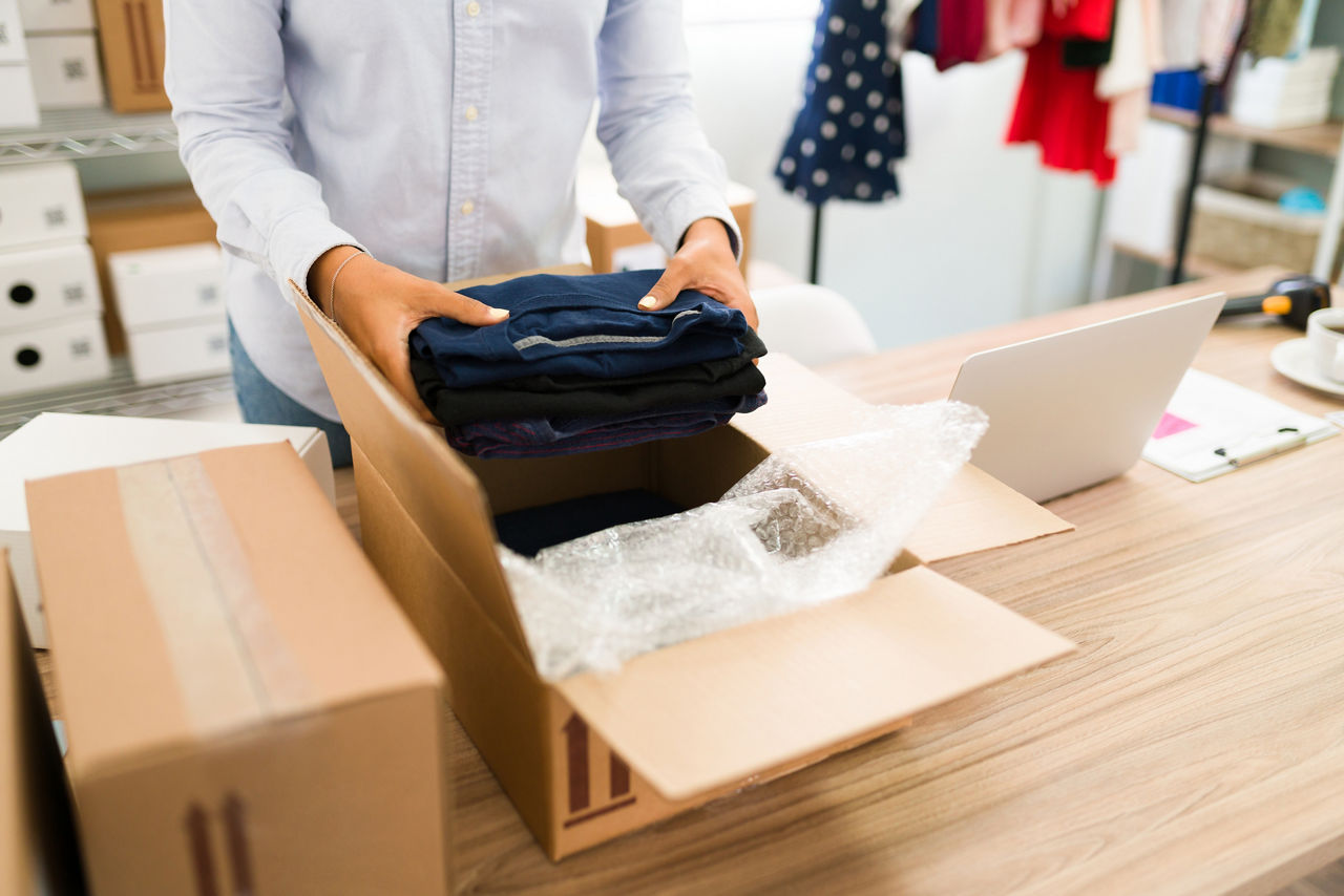 Ready to ship a package from my online fashion shop. Young woman putting t-shirts on a box to send in the mail to a customer