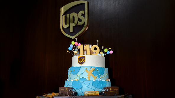 A multi-tiered UPS-themed cake is displayed against a dark wooden wall with a large UPS shield emblem. The cake features a white top tier with the number 119 and colorful balloon decorations, and a blue lower tier with delivery route graphics. Small UPS delivery truck models are placed around the base, along with a plaque reading "Happy 119th Anniversary UPS." The scene suggests a festive corporate celebration with bright colors contrasting the dark background.