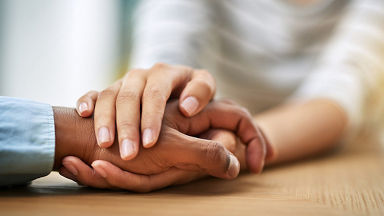 A close-up image of two hands gently holding each other on a wooden surface. The setting suggests a moment of comfort or emotional support. The hands belong to individuals of different skin tones, emphasizing connection and empathy.
