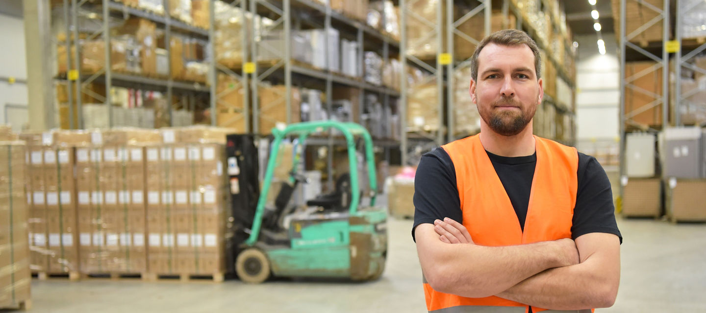 Male in a vest crossing his arms with forklift, pallet and warehouse racks in the background