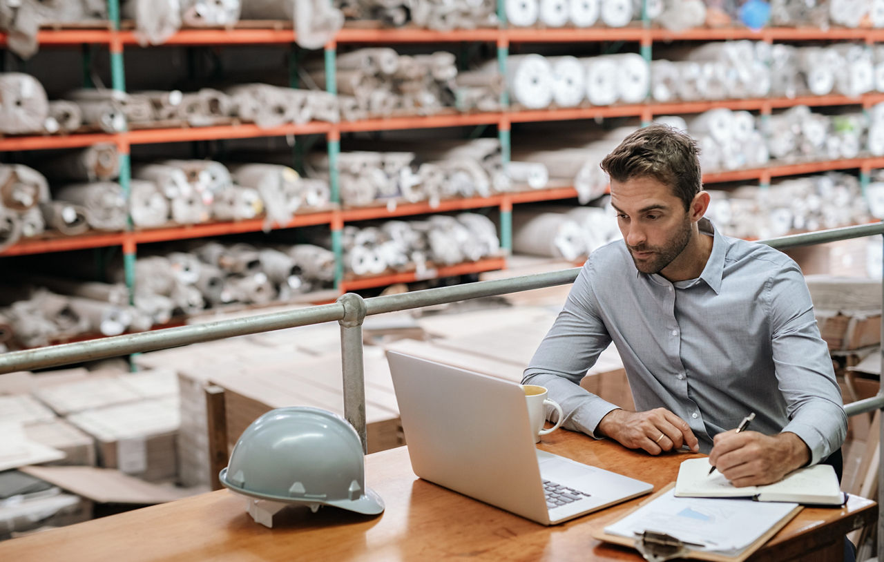 Manager checking inventory and online orders on a laptop while sitting at a desk in a carpet warehouse with shelves of stock in the background; Shutterstock ID 1414684886; purchase_order: New Web Images; job: ; client: ; other: 