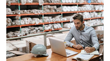 Manager checking inventory and online orders on a laptop while sitting at a desk in a carpet warehouse with shelves of stock in the background; Shutterstock ID 1414684886; purchase_order: New Web Images; job: ; client: ; other: 