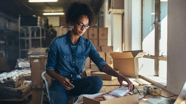 Female sitting on a stool and is preparing an order surrounded by shipping supplies 