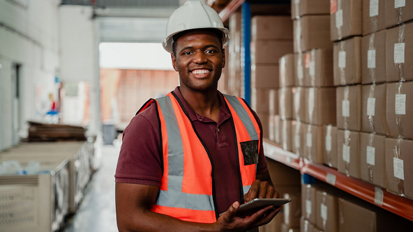 Male in a safety vest and hard hat holding a tablet in a warehouse 