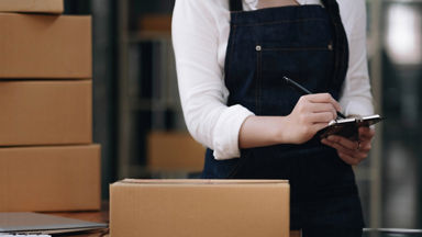Female wearing an apron and writing on a clipboard with a box in front of her