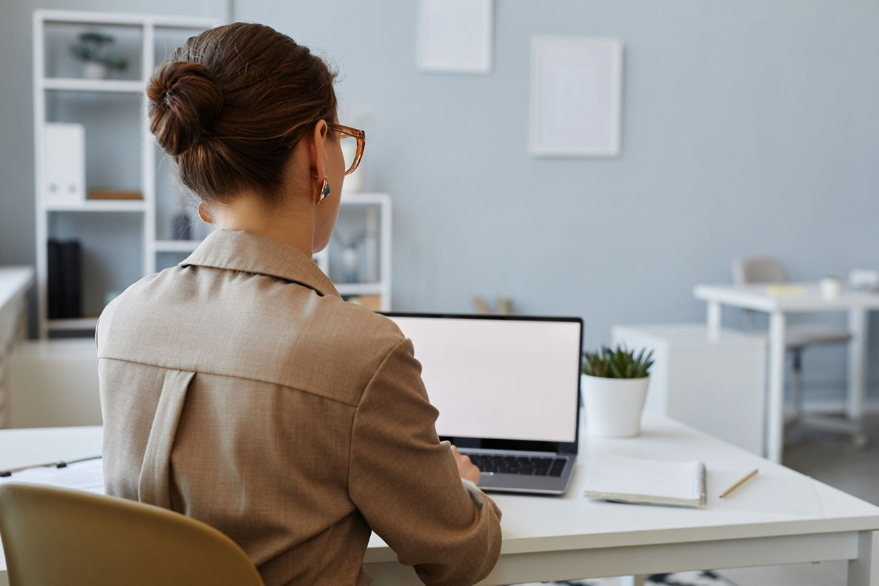 Back view of young businesswoman using laptop with blank screen mockup at workplace in office, copy space