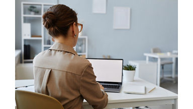 Back view of young businesswoman using laptop with blank screen mockup at workplace in office, copy space