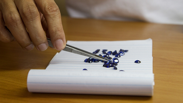 Close up of hands sorting blue gemstones with tweezers on lined paper